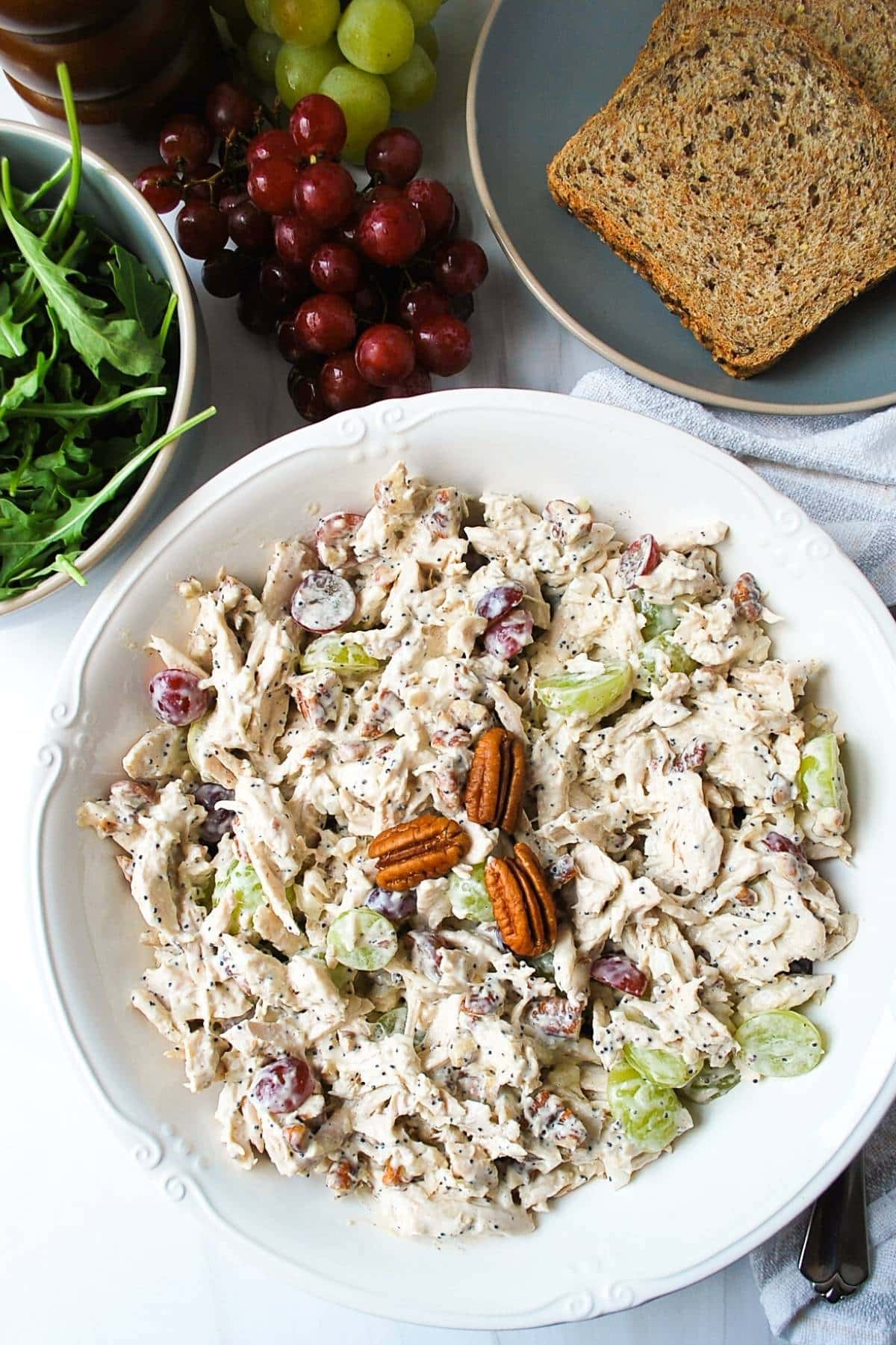 overhead photo of a serving bowl of California chicken salad topped with a few whole pecans with grapes, greens, and toasted bread in the background