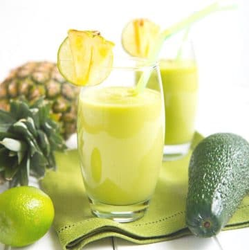 two green smoothies in a glass with limes, avocado, and a pineapple