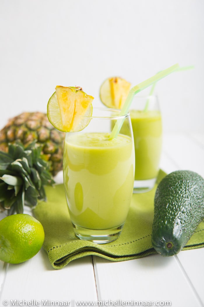 two pineapple avocado smoothies in a glass with an avocado and fresh pineapple next to the glasses