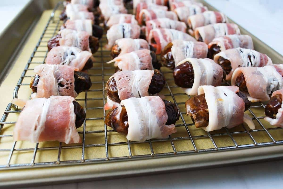 dates stuffed with an almond wrapped in bacon on a wire rack over a parchment lined baking sheet ready to bake