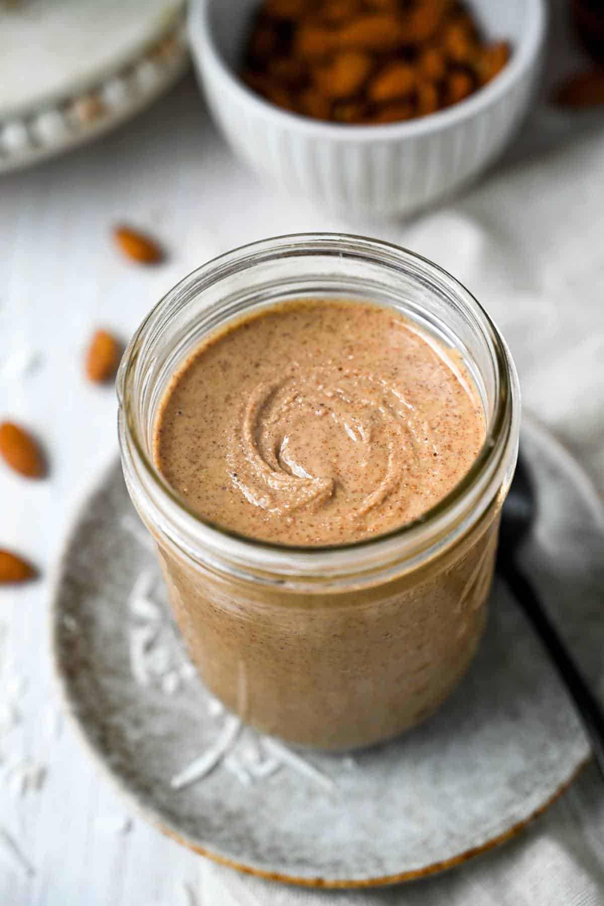 a jar of almond butter on a plate with a spoon with nuts and coconut