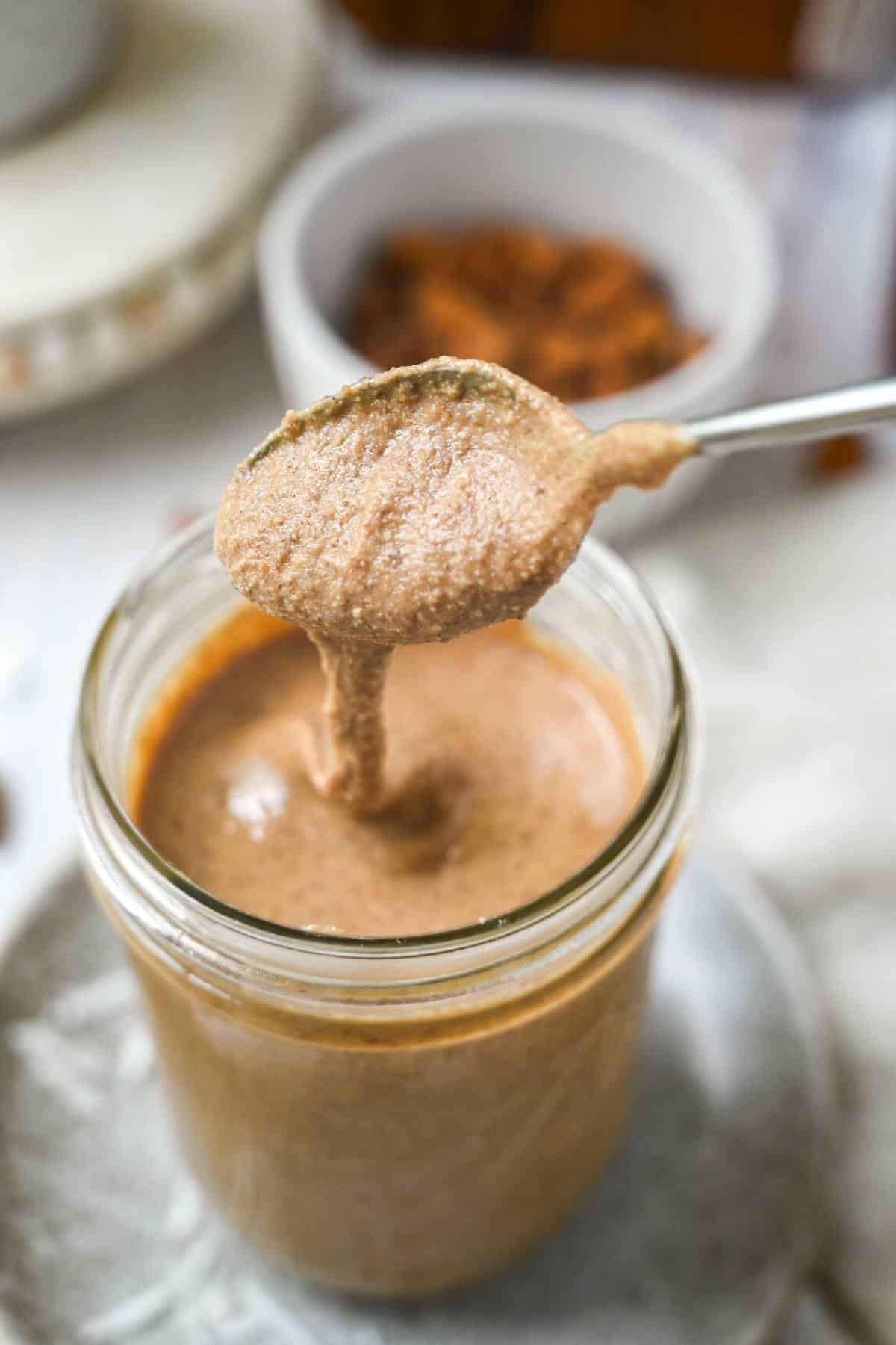 a spoonful of almond butter over a jar of almond butter with a dish of almonds behind it