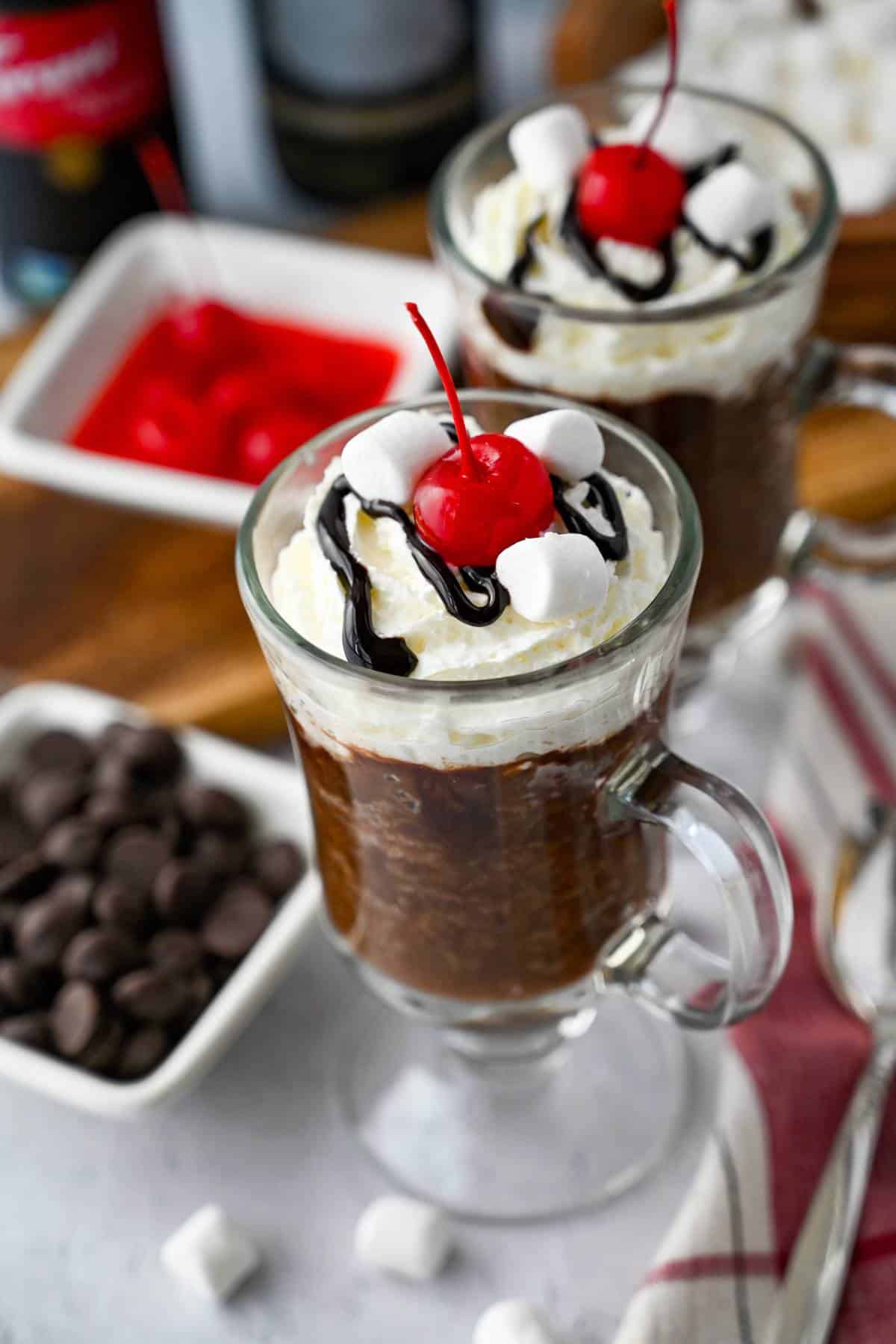 two mug cakes made with hot cocoa mix topped with whipped cream, chocolate sauce, and cherries with a bowl of chocolate chips and cherries next to them