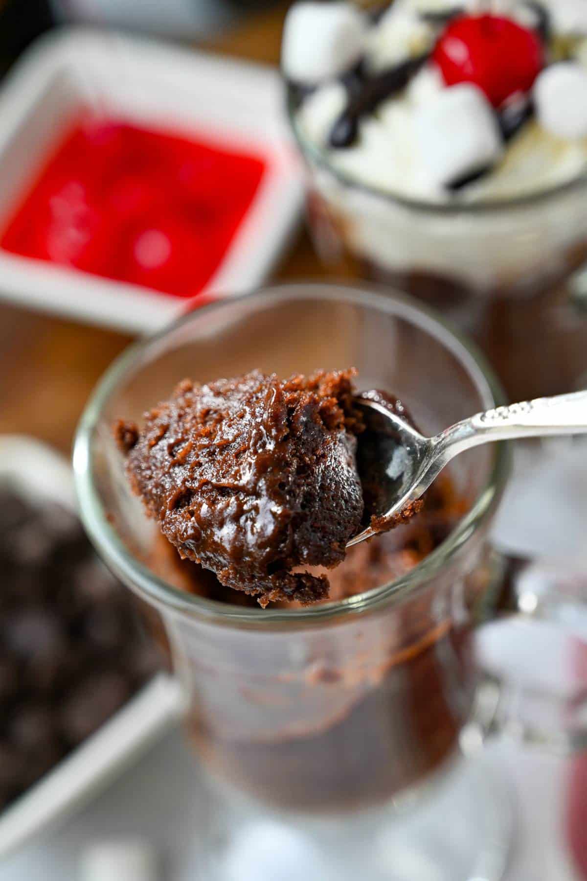 a spoonful of gluten free chocolate mug cake with a bowl of cherries and mug cake with toppings behind it
