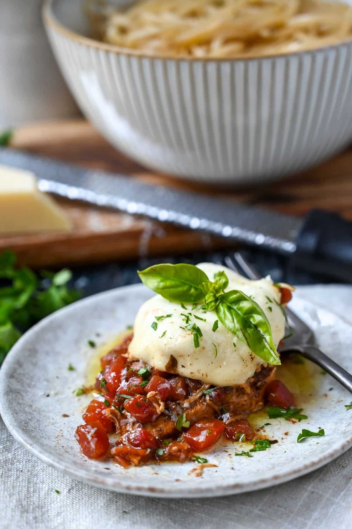 Italian chicken topped with melted mozzarella on a plate with a bowl of pasta behind it