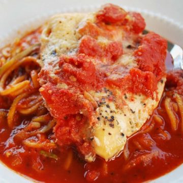 close up photo of a cooked chicken breast topped with tomatoes, cheese, and spices over a bed of pasta with marinara sauce