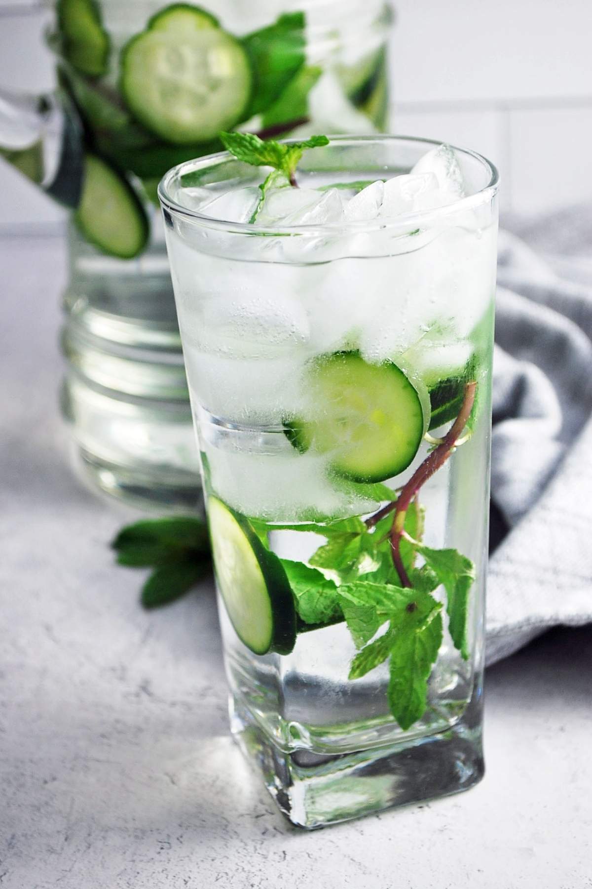 glass of water with sliced cucumbers and a mint sprig inside with ice and a pitcher of infused water in the background