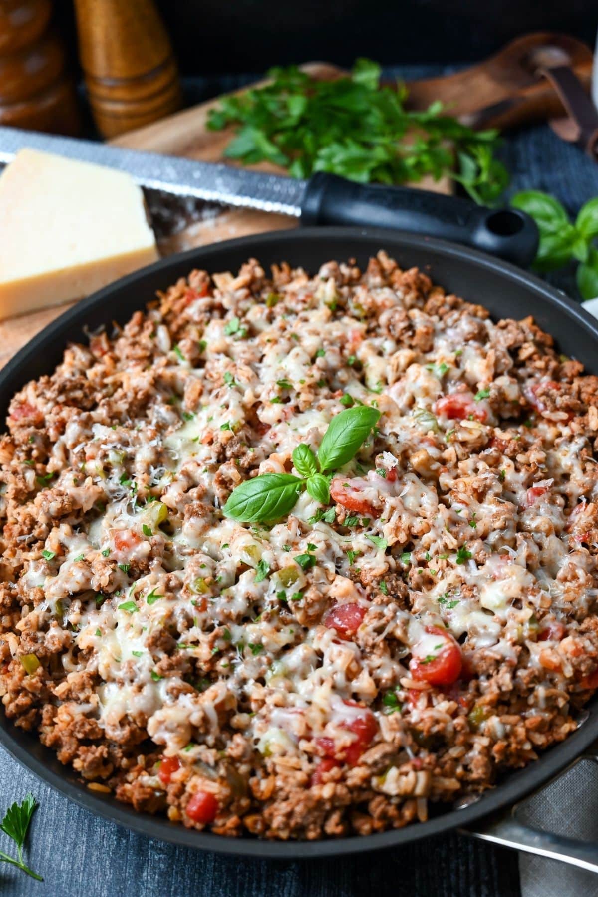 a cooked Italian beef skillet with a sprig of fresh basil on top with a cutting board with a block of parmesan and fresh parsley behind it