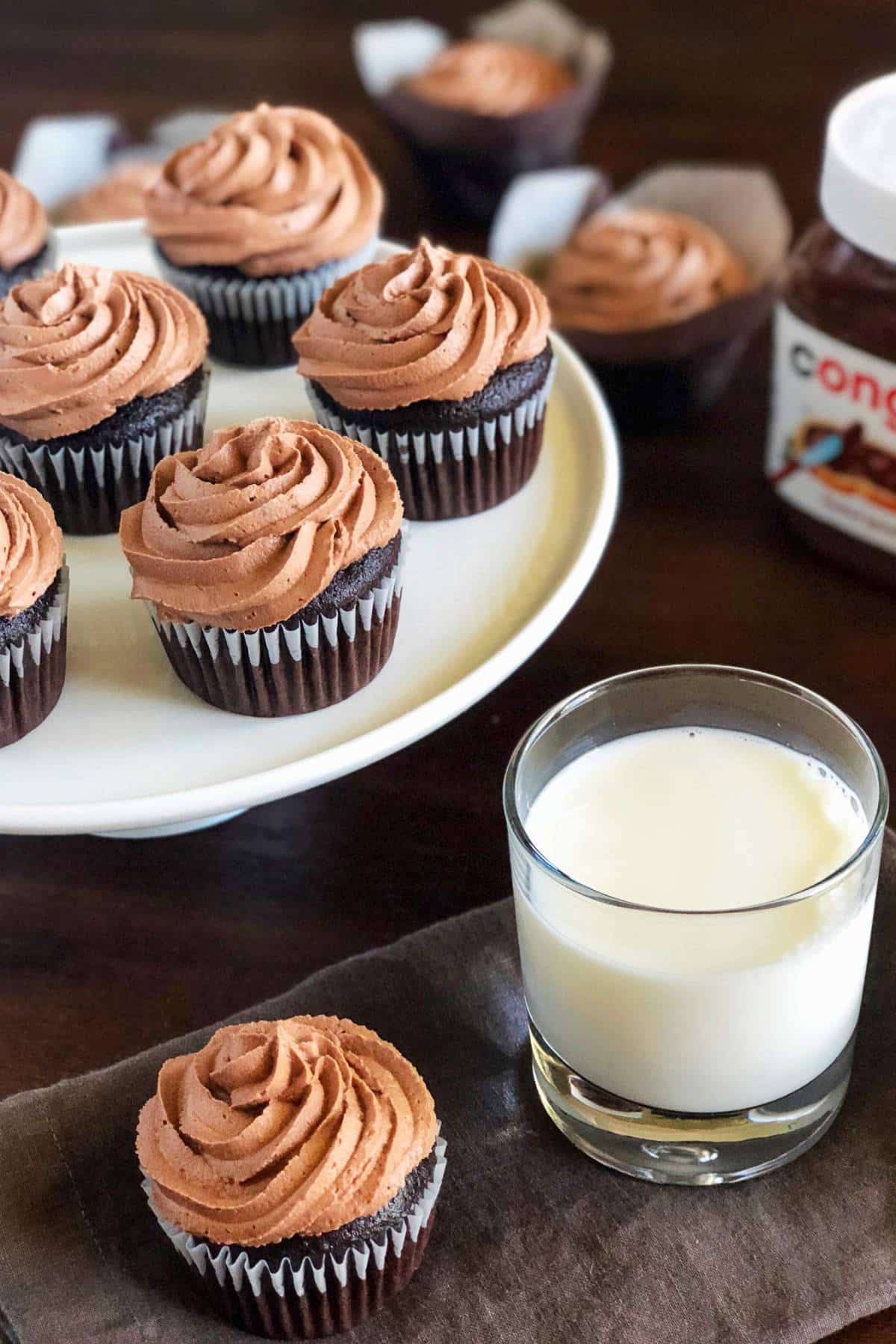 a platter of chocolate cupcakes with light brown chocolate hazelnut frosting next to a brown napkin with a cupcake and a glass of milk