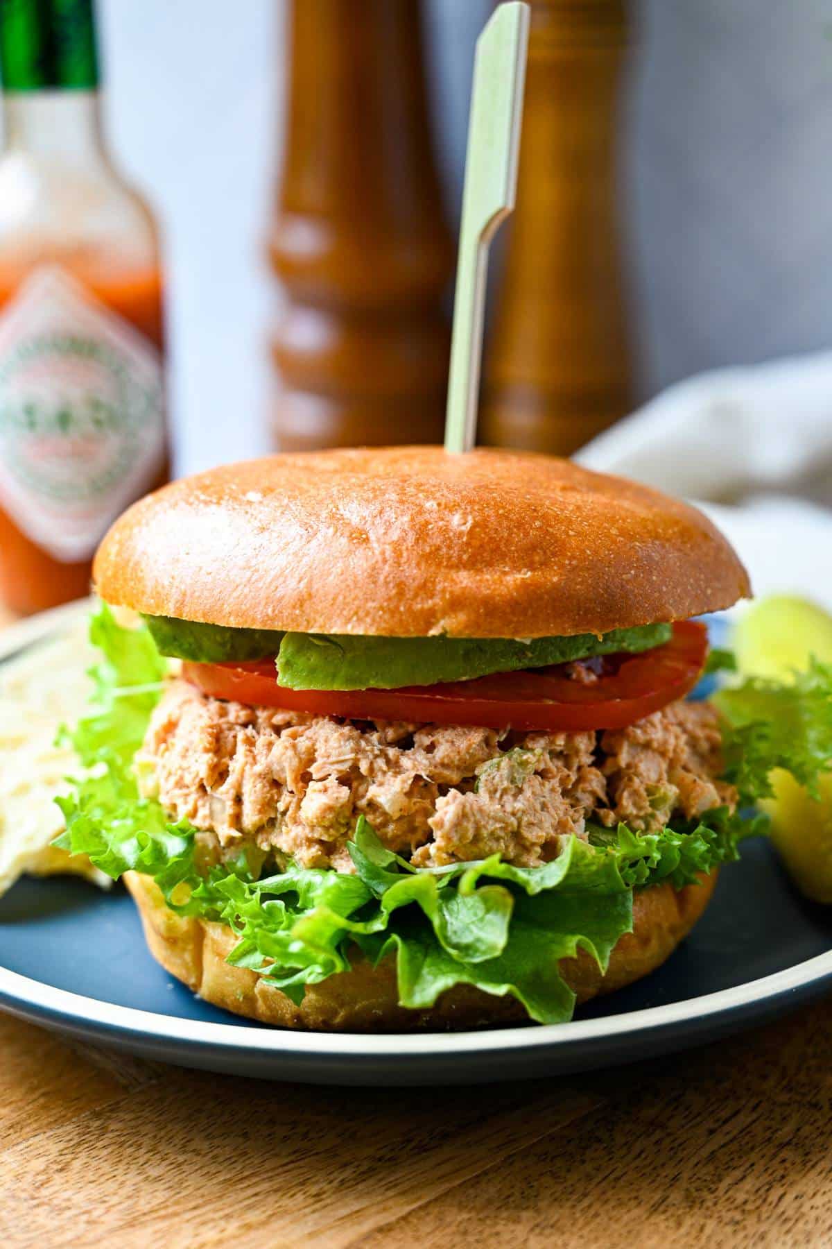 spicy tuna salad on a brioche bun with lettuce, tomato, and avocado with a pickle and chips on a plate