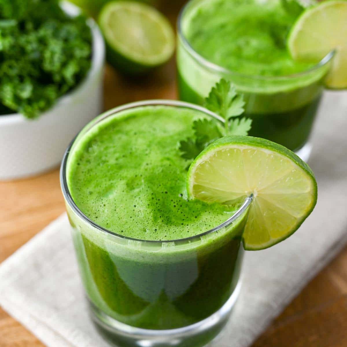 two glasses of green juice with a bowl of kale behind them