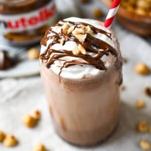 a hazelnut protein shake in a glass topped with whipped cream drizzled nutella and a straw