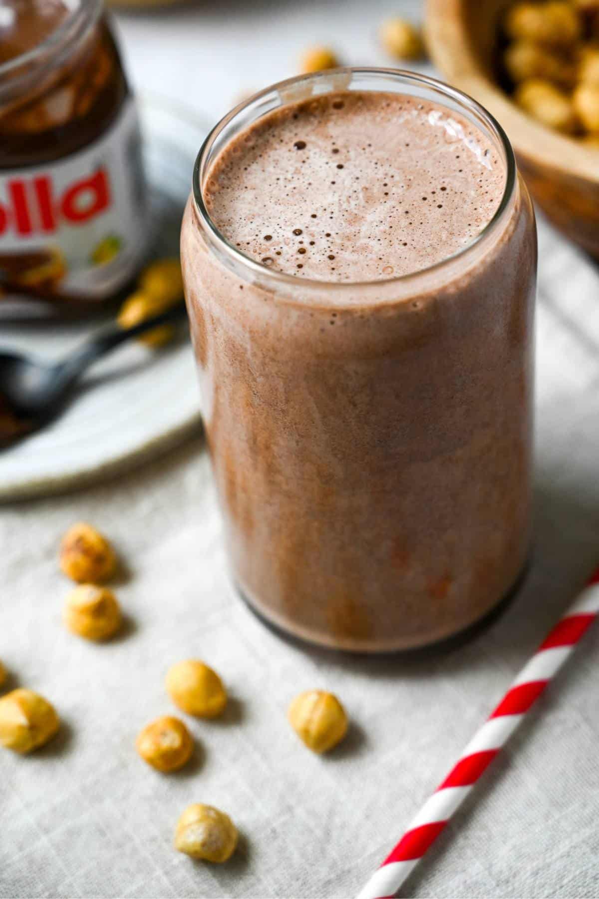 a chocolate hazelnut shake in a glass with hazelnuts and a straw next to it