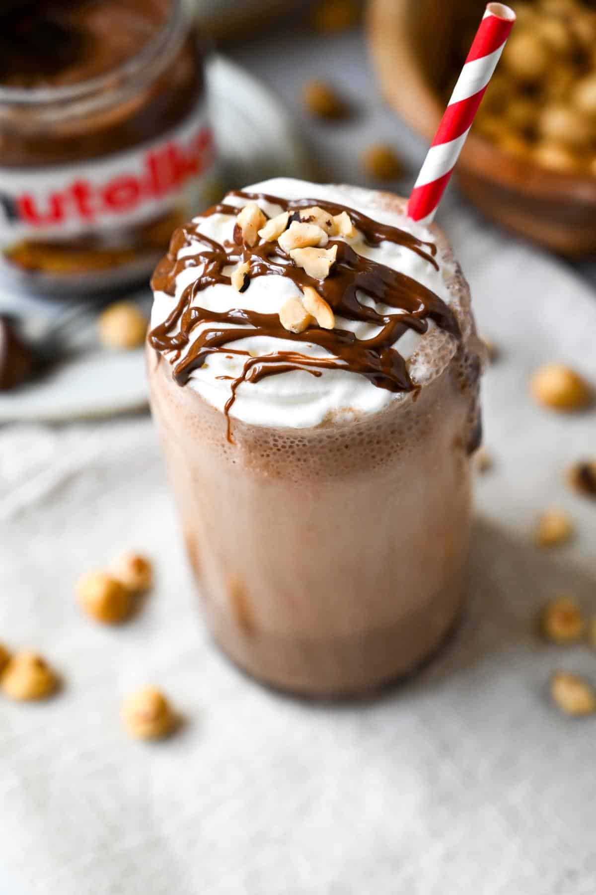a chocolate hazelnut protein shake in a glass topped with whipped cream, nutella drizzle and chopped hazelnuts with a jar of nutella and a bowl of hazelnuts 