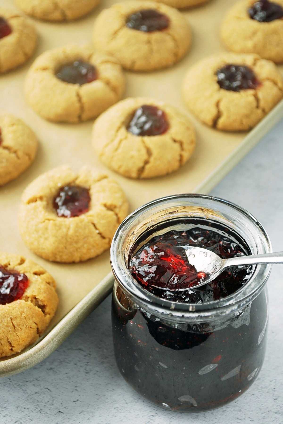 jar of grape jam with a spoon with baking sheet of cookies filled with jam in the background