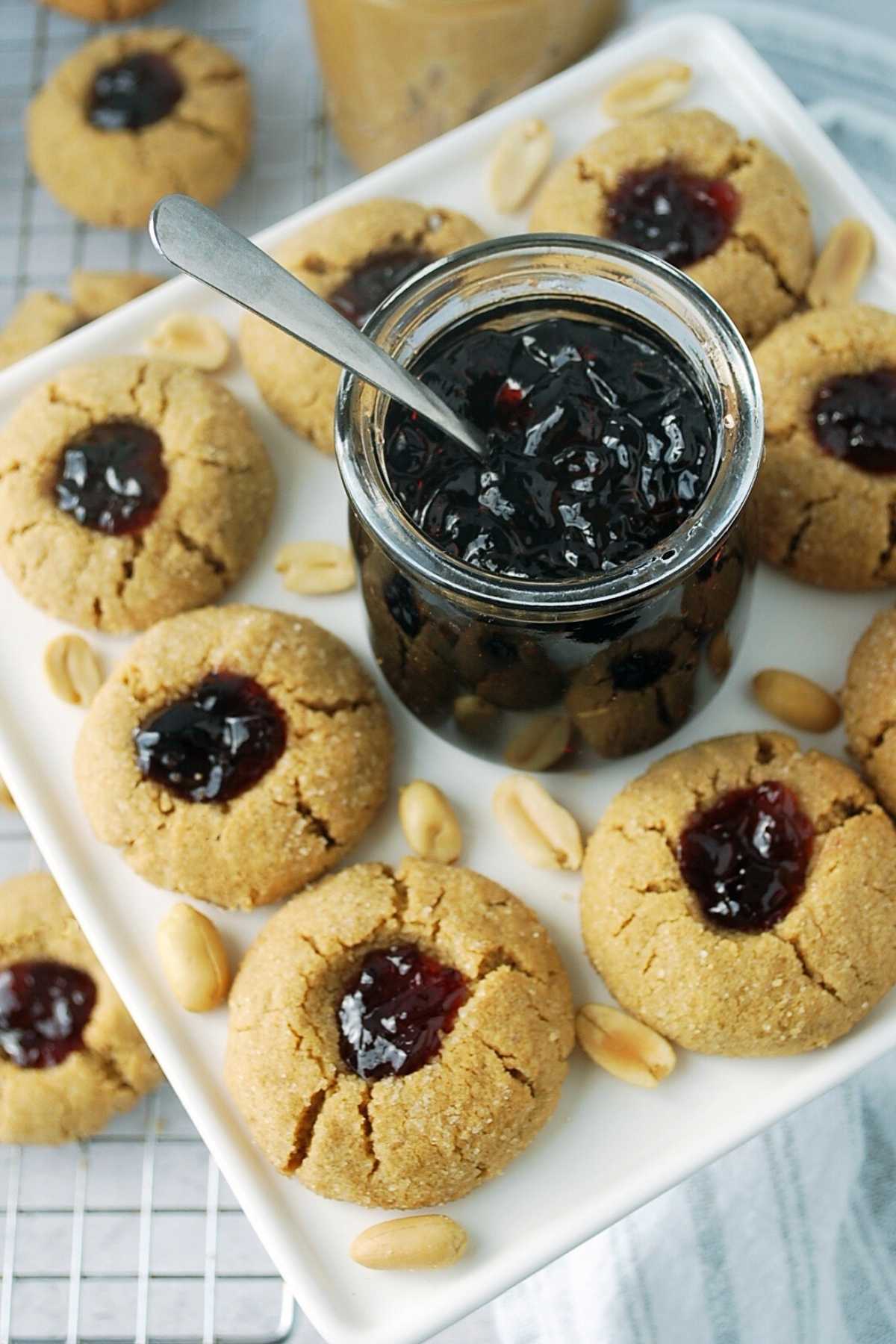 overhead photo of peanut butter thumbprint cookies with a jar of jam in the middle and peanuts sprinkled around the platter and cooling rack of cookies underneath 