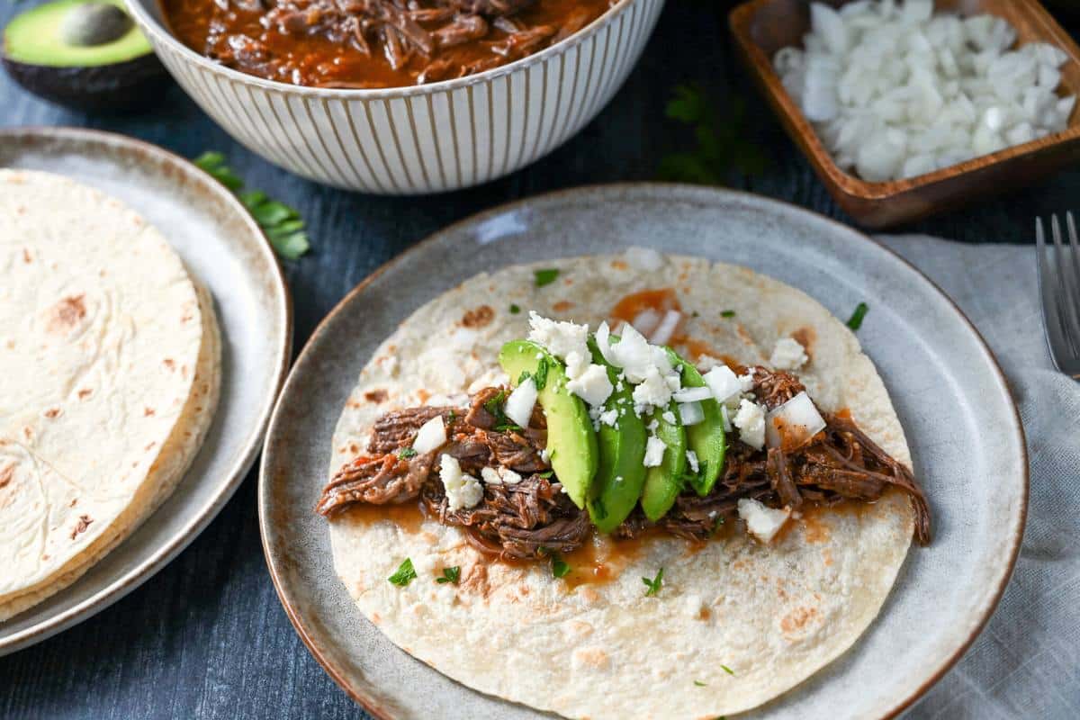 a shredded beef taco with a plate of flour tortillas, avocado, onion, and a bowl of shredded beef