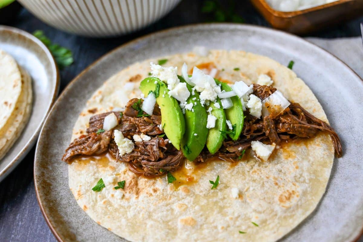 a plate with a shredded beef taco with avocado, onion, and cheese topping