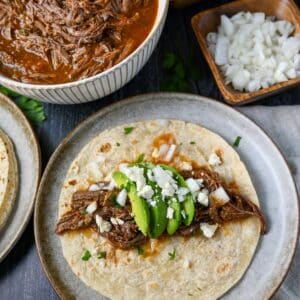 overhead photo of a plate with a tortilla topped with southwest pot roast, avocado slices, onion, and queso fresco cheese with a bowl of beef roast and onions in the background