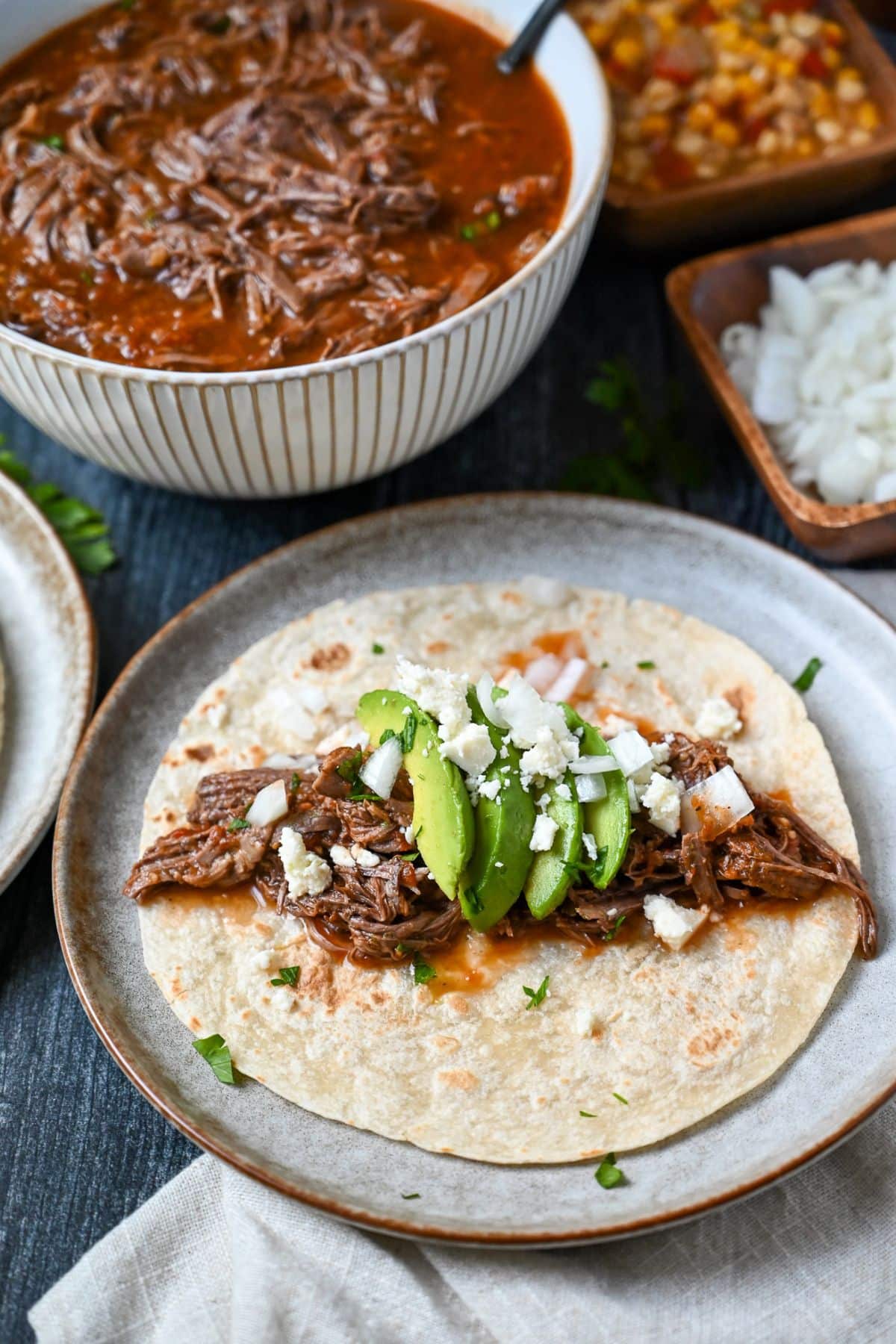 a shredded beef taco with avocado, onions, and cheese with a bowl of shredded beef