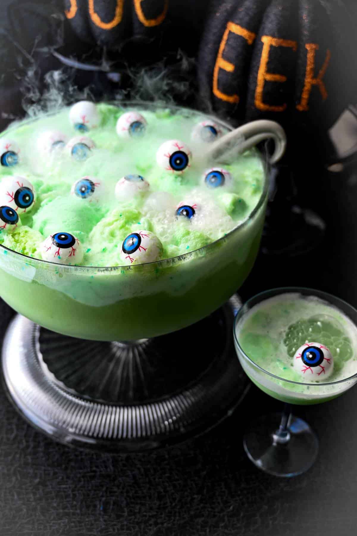 a punch bowl of green witches brew punch with floating eyeballs and dry ice for a smokey effect with a martini glass of bubbling punch next to it