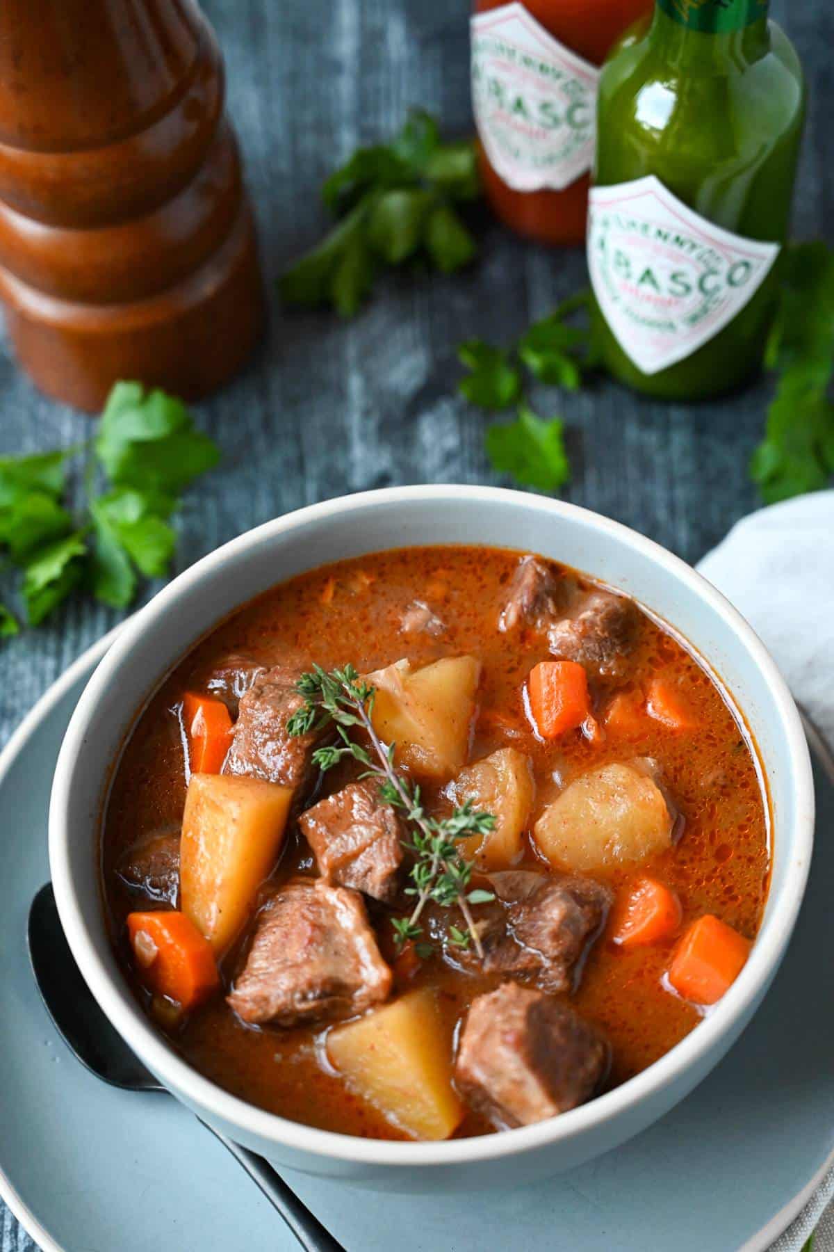 beef stew in a gray bowl with a spoon with a peppermill, fresh herbs and seasonings in the background