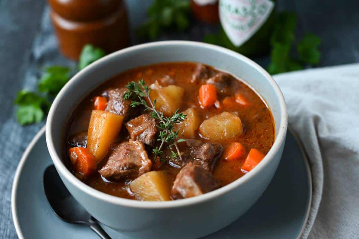 a bowl of beef stew with fresh herbs and Tabasco bottle in the background