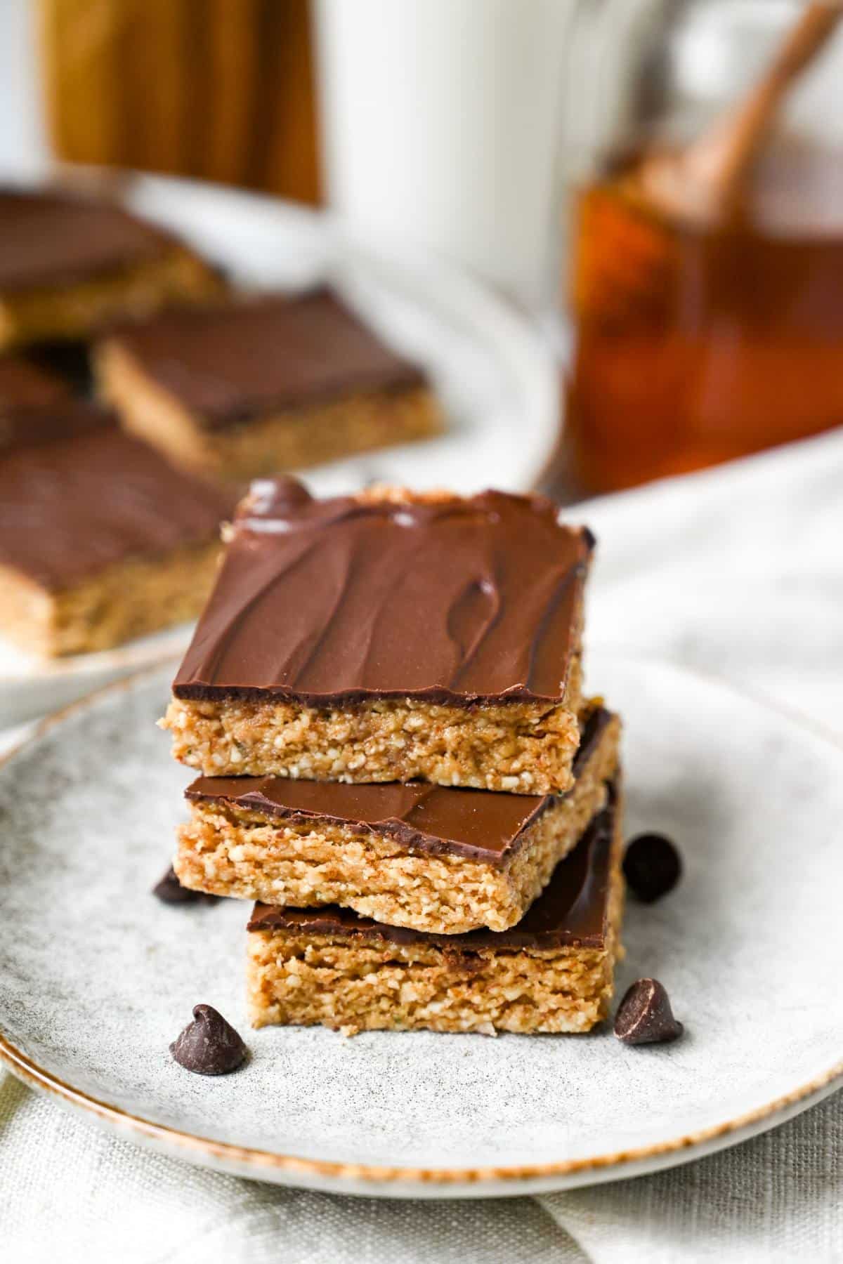 3 dessert bars stacked on a plate with a jar of honey, glass of milk, and plate of bars behind it