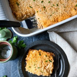 a pan of hashbrown casserole with a serving on a black plate with fresh herbs next to it
