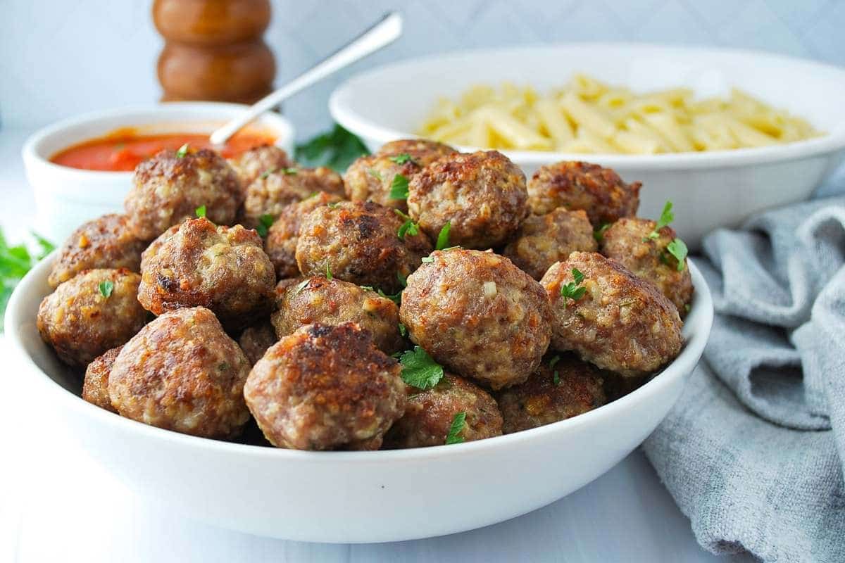 gluten-free meatballs in a white bowl with a bowl of marinara and pasta in the background