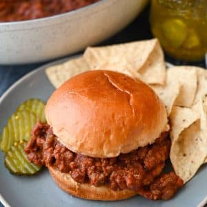 a sloppy joe on a plate with pickle slices and tortilla chips