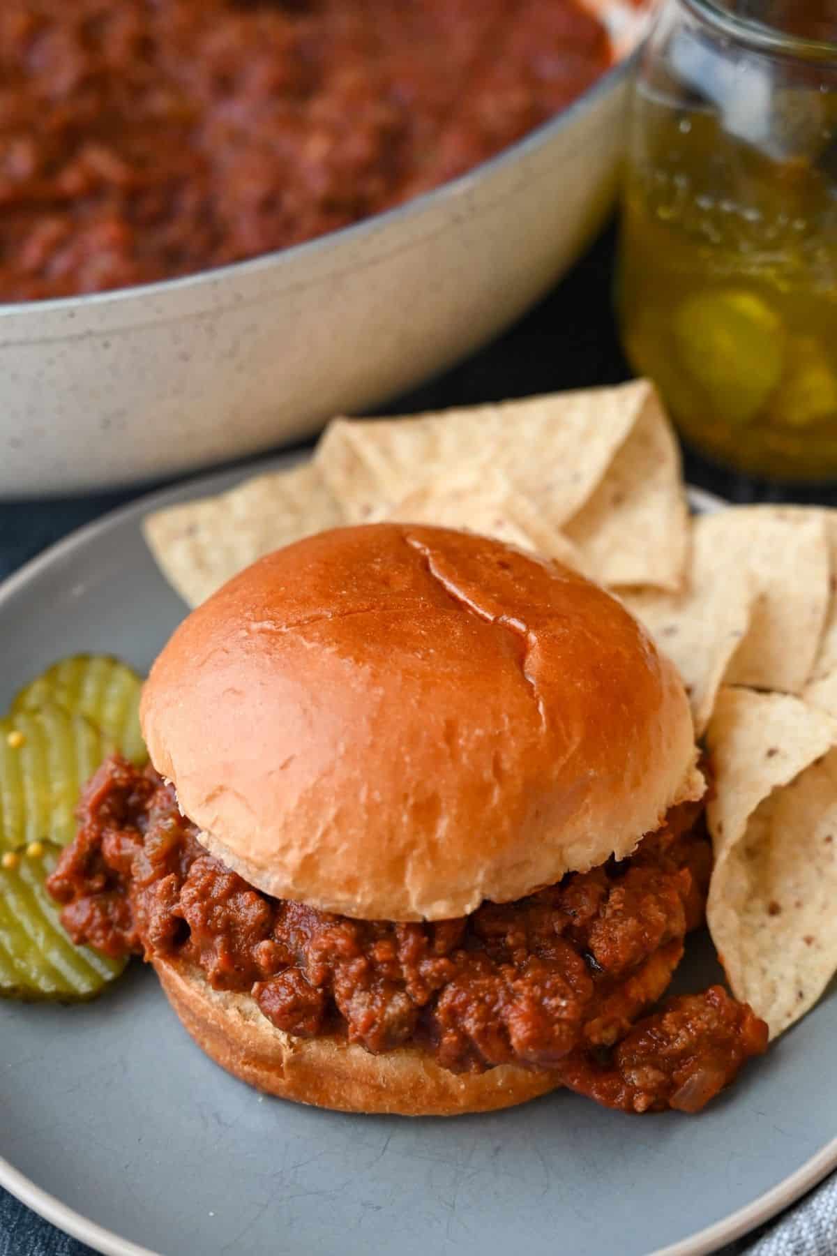 looking down at a plate with a sloppy joe with pickles and chips and a jar of pickles and skillet of venison sloppy joe meat behind it