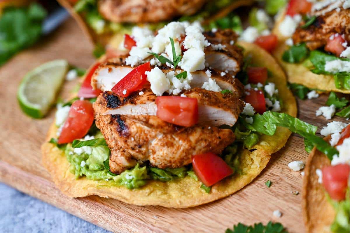 a tostada with avocado and grilled chicken loaded with toppings on a wood serving board
