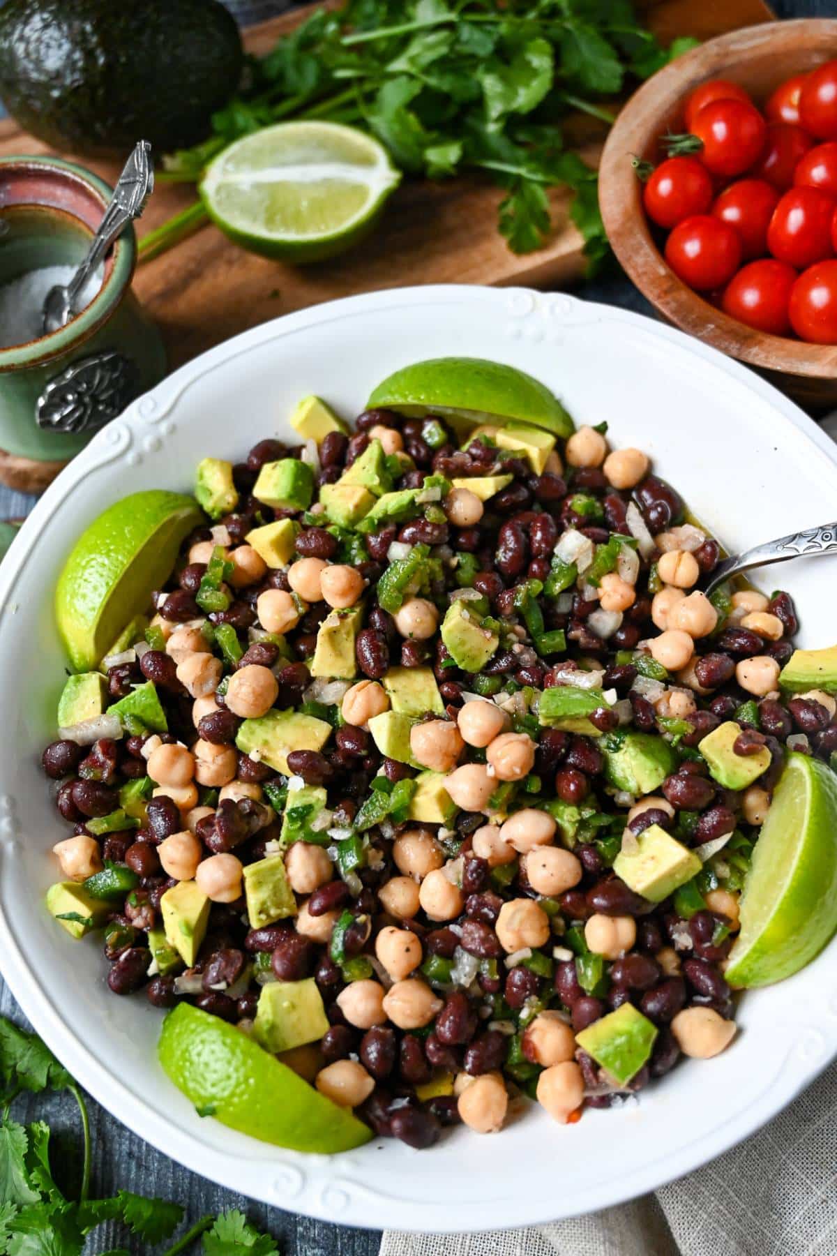 a bowl of bean salad with limes, avocado and cilantro with a spoon and fresh cilantro and vegetables behind it