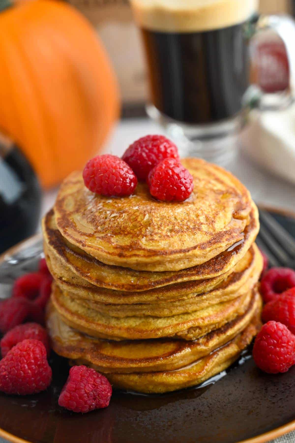 close up of a large stack of pumpkin pancakes with fresh raspberries and a cup of coffee next to it