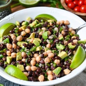 close up of a serving bowl of black bean and chickpea salad with avocado and lime slices