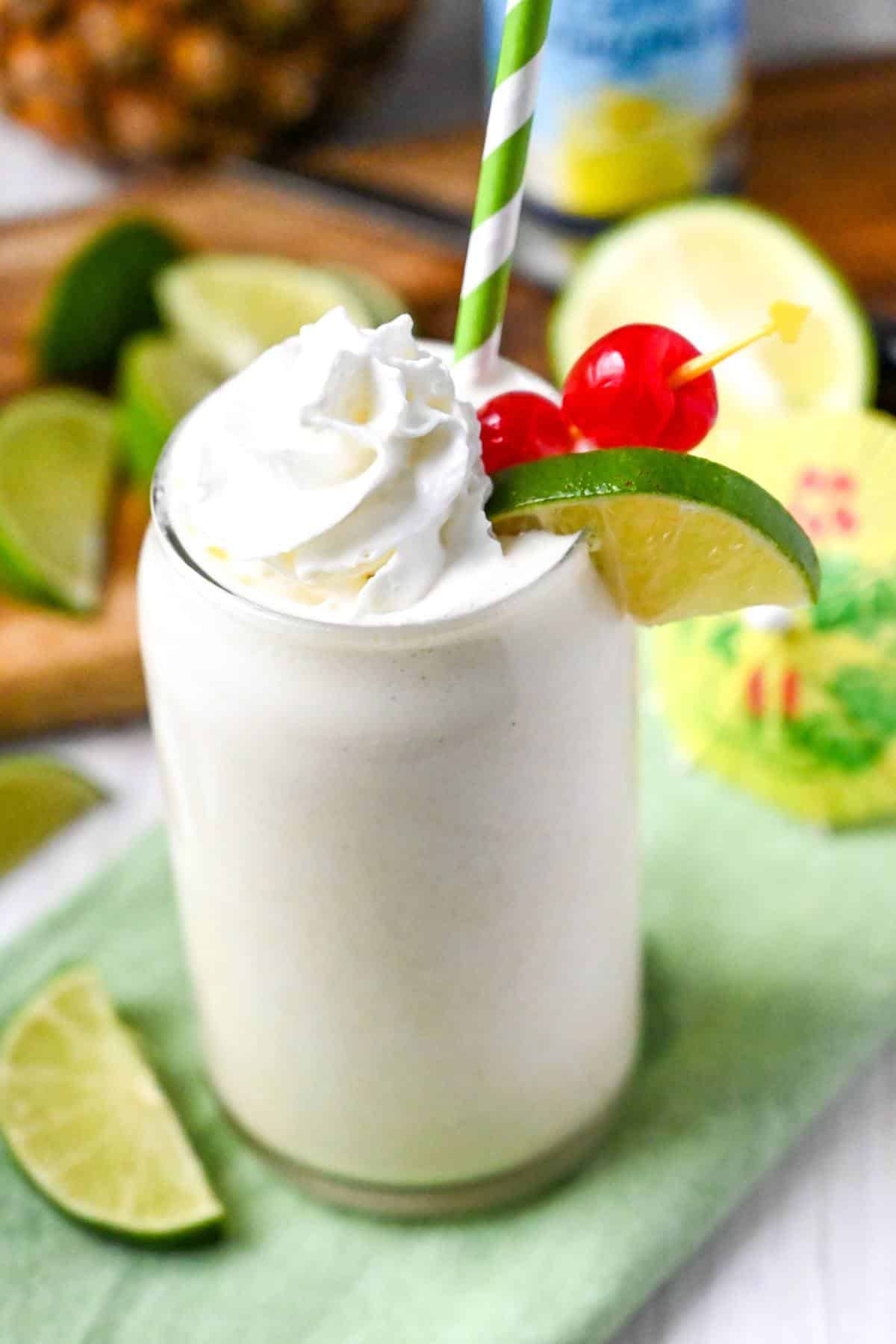 a pineapple banana protein shake with a green striped straw and a cutting board of limes, can of pineapple juice and a whole pineapple in the background