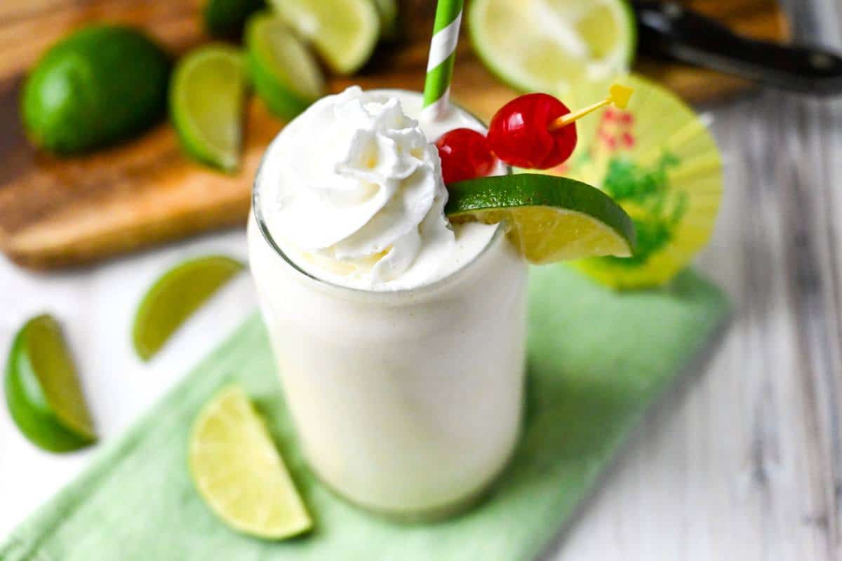 a protein shake in a glass with lime, whipped cream and cherries with a straw with a cutting board with lime wedges