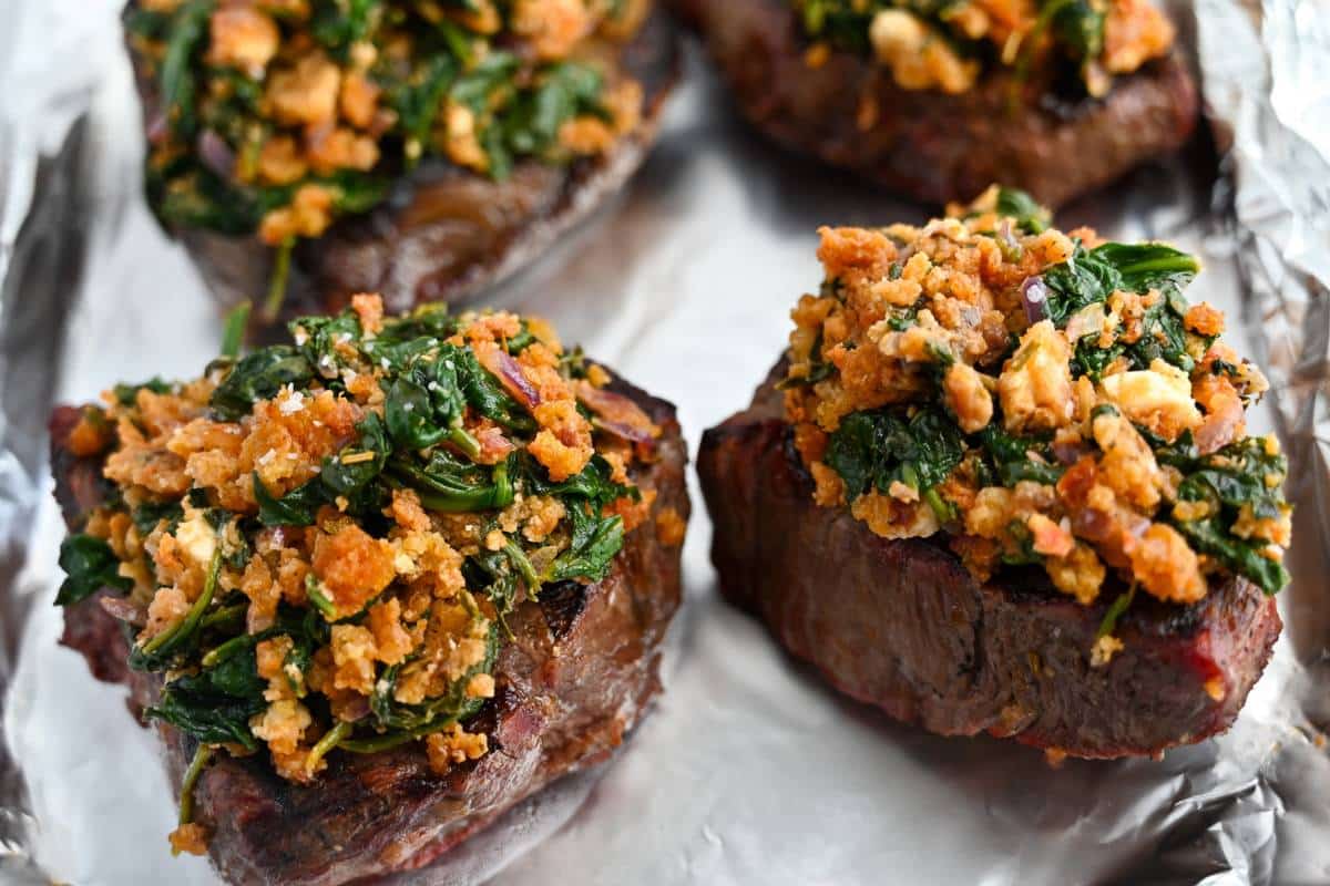 a feta spinach sundried tomato topping on top of grilled steaks on a foil-lined baking sheet