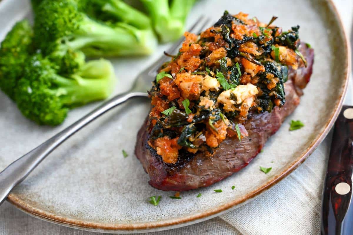 a steak on a plate with a broiled topping with a knife, fork, and napkin