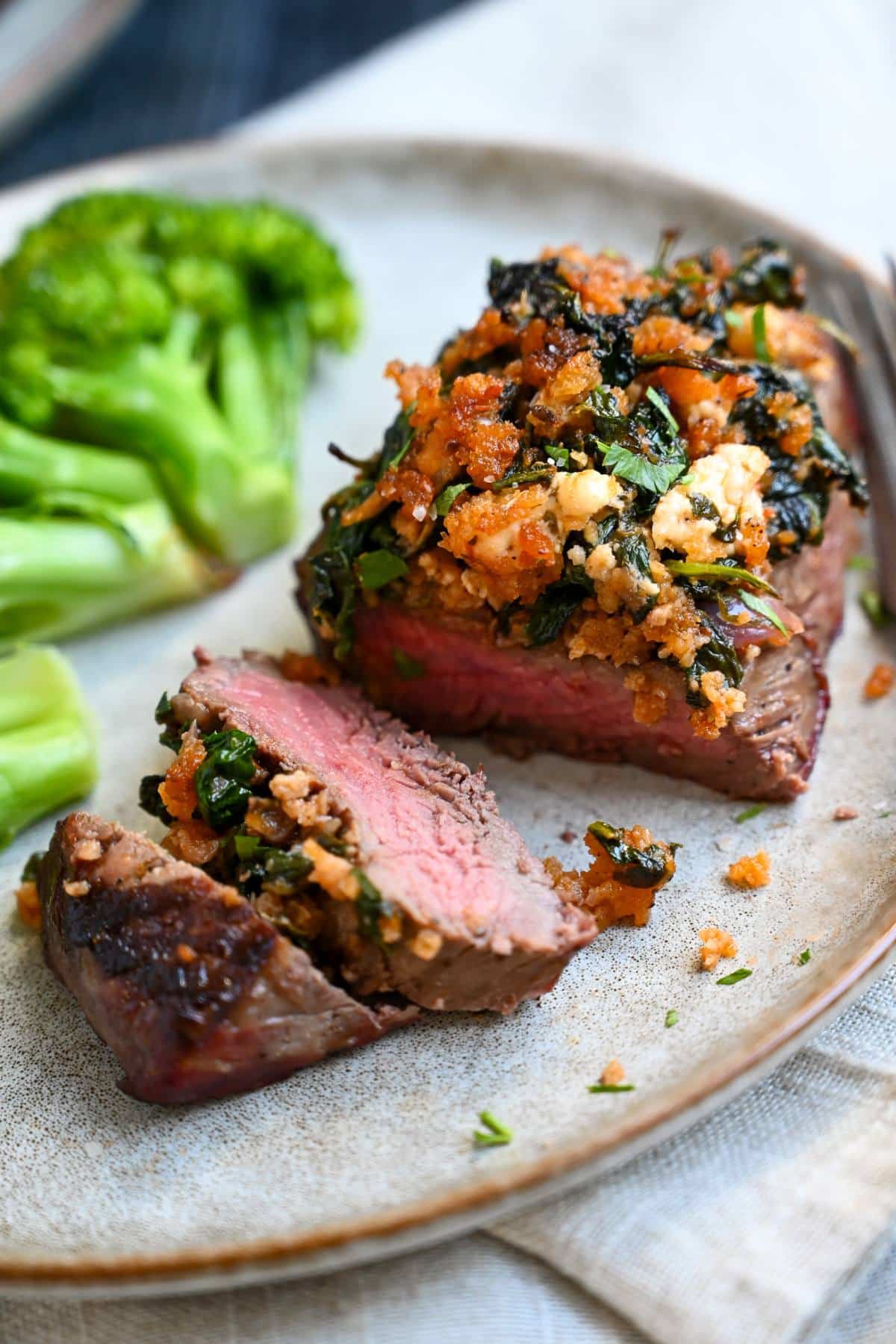 a sliced sirloin steak topped with a Mediterranean inspired topping on a plate with broccoli