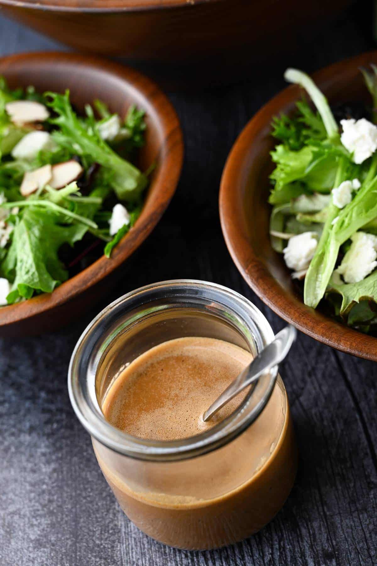 a jar of salad dressing with a spoon in front of two salad bowls