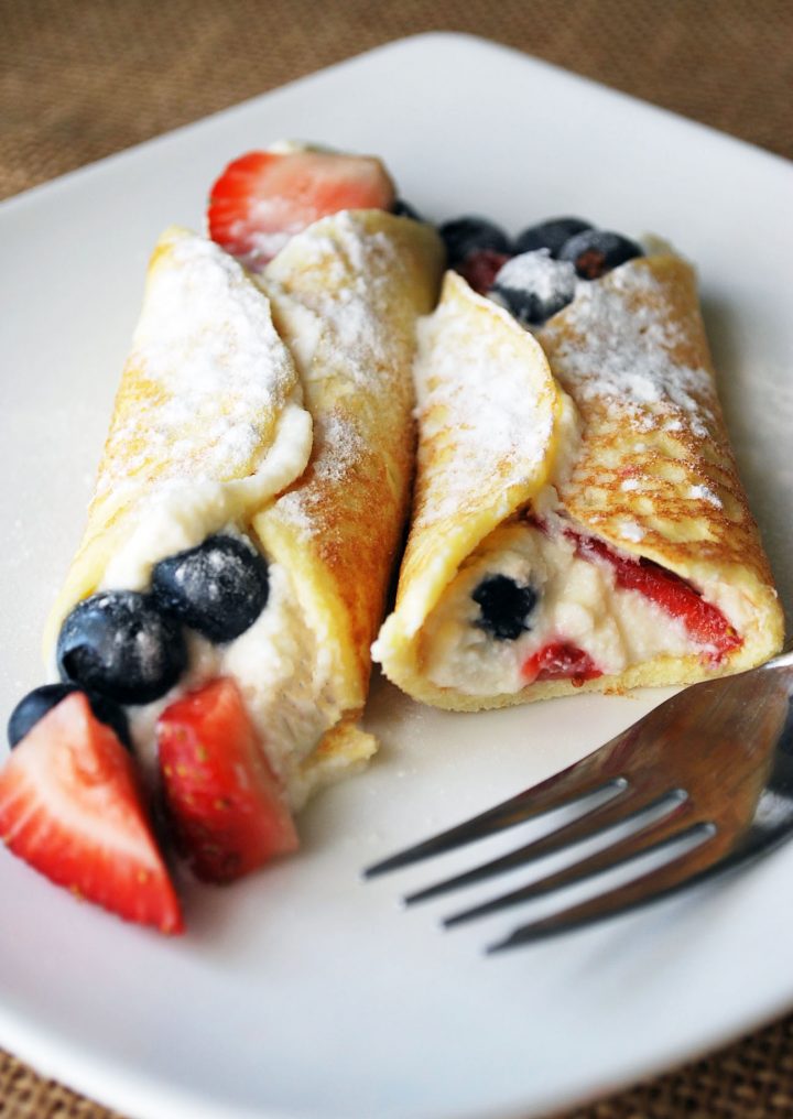 half eaten stuffed crepe with a whole stuffed crepe with berries and powdered sugar on a plate