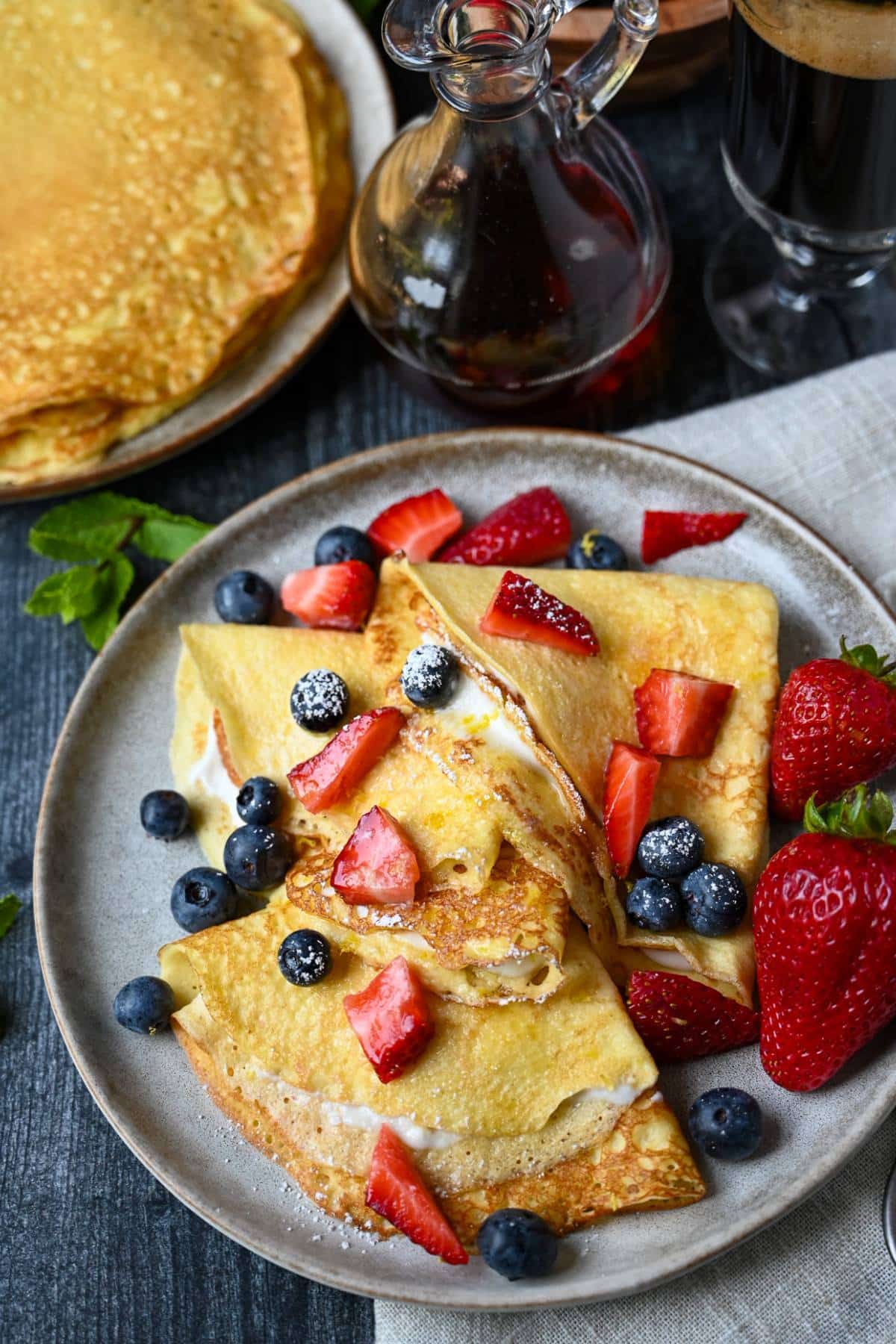 looking down at a plate of folded stuffed crepes with berries and a plate of plain crepes with a jar of syrup