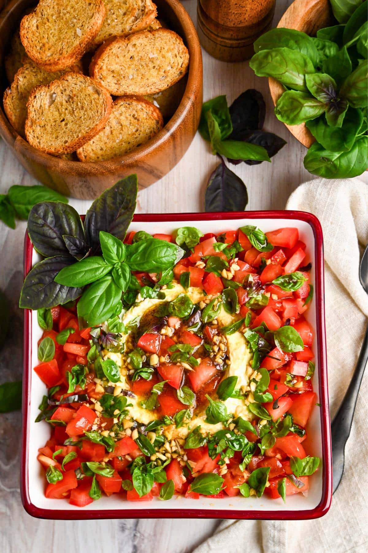 a caprese cheese ball in a bowl with tomatoes, basil, toast points and a bowl of fresh basil