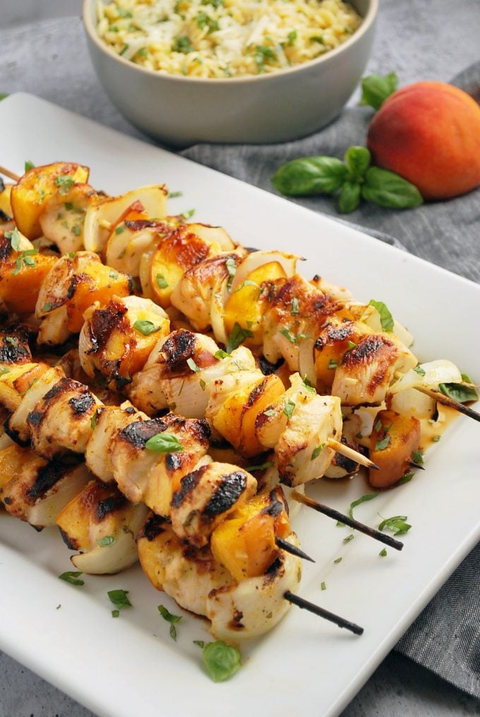 chicken and peach kabobs on a platter garnished with basil with peach and bowl of orzo in the background