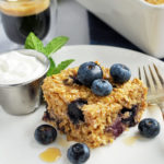 Slice of blueberry baked oatmeal on a plate topped with fresh blueberries and a side of yogurt