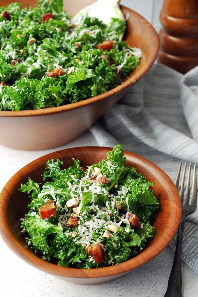 close up of kale salad in a small wood bowl with a serving bowl of salad and a peppermill in the background