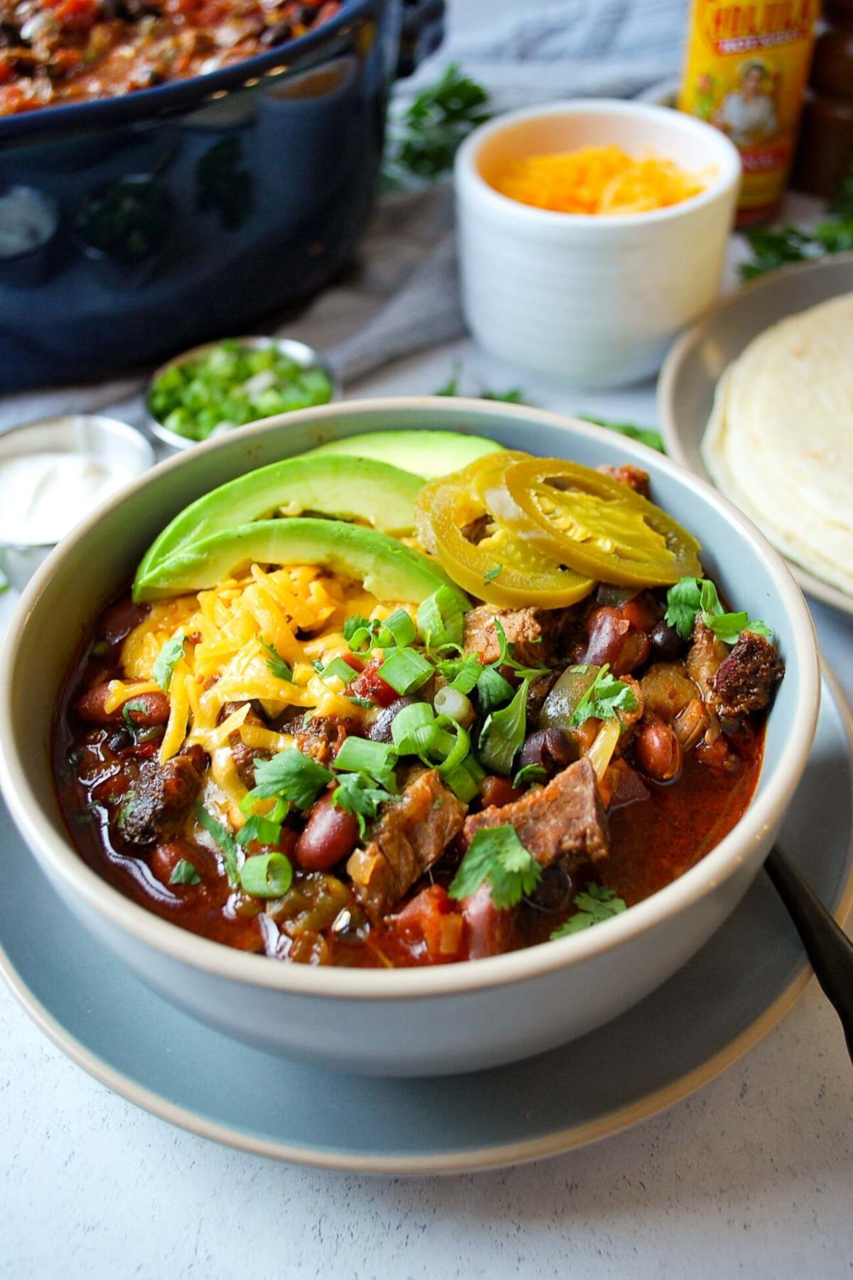 close up of a bowl of brisket chili topped with sliced avocado, cheese, and green onions