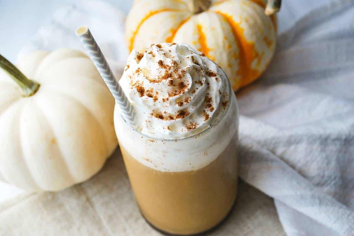 an iced pumpkin spice latte with a straw and whipped cream next to two mini pumpkins and a napkin